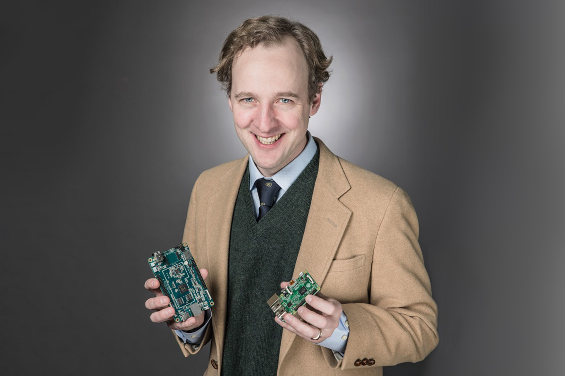 portrait of a smiling Anatole von Lilienfeld, wearing a tan blazer and dark grey vest and tie, holding a circuit board in each hand