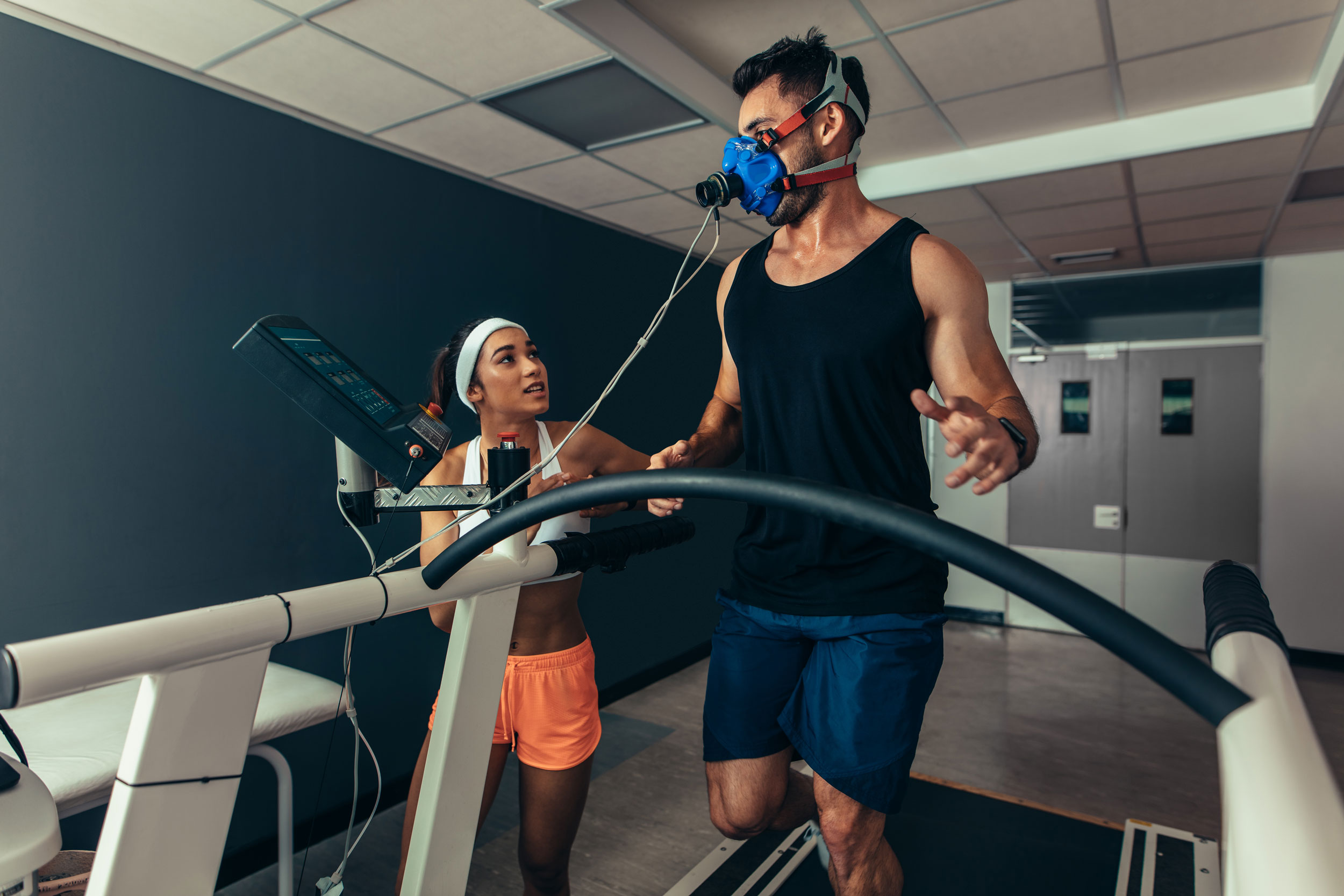 Athlete wearing monitoring equipment, running on a treadmill, accompanied by another athlete