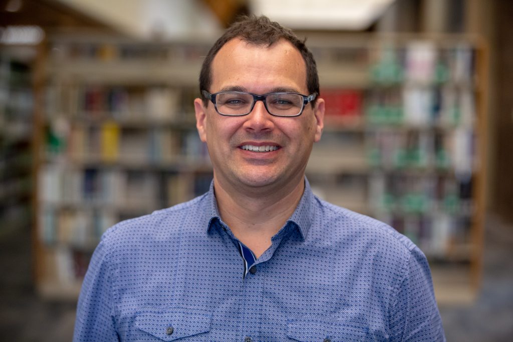 Steven Hermans wearing a blue shirt and glasses, in the background is a library bookshelf