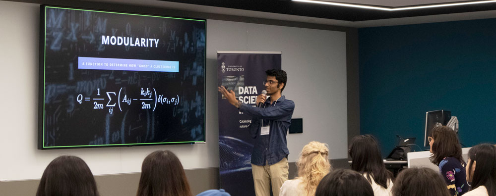 A young South Asian student holding a mic and gesturing to a screen with the title" Modularity" presenting to a classroom of students