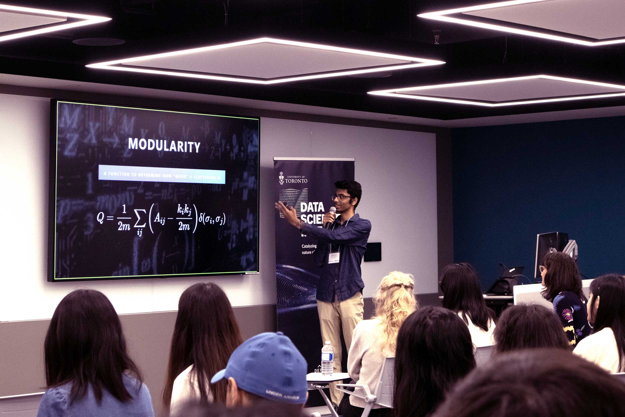 A young South Asian student holding a mic and gesturing to a screen with the title" Modularity" presenting to a classroom of students