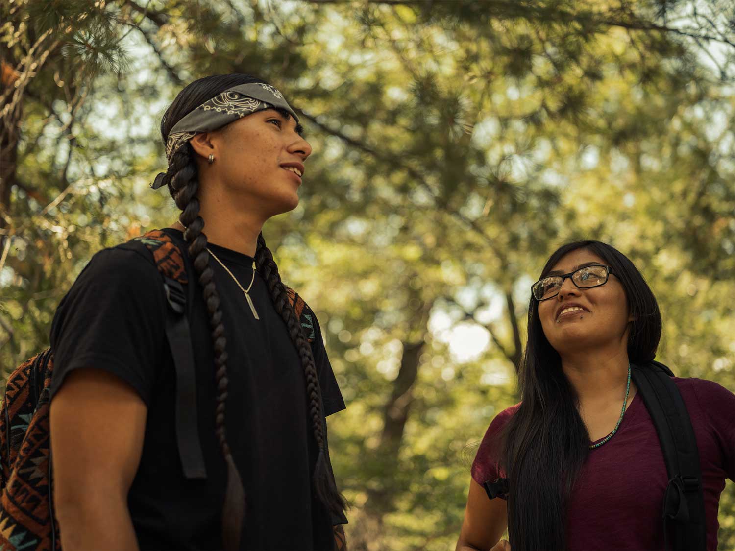 Two long-haired Indigenous students, one with braids, the other with glasses, with trees in the background